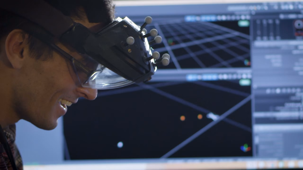 Close profile of a developer wearing the AR headset, smiling, a 3D tracking grid on the monitor behind him